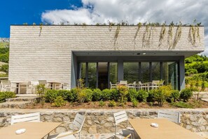A modern building with a brick facade, large glass windows, outdoor seating, and a stone retaining wall. - Odat n`Bogë | Room 3 - Mali | Breakfast Included by PikHost (Bogë)