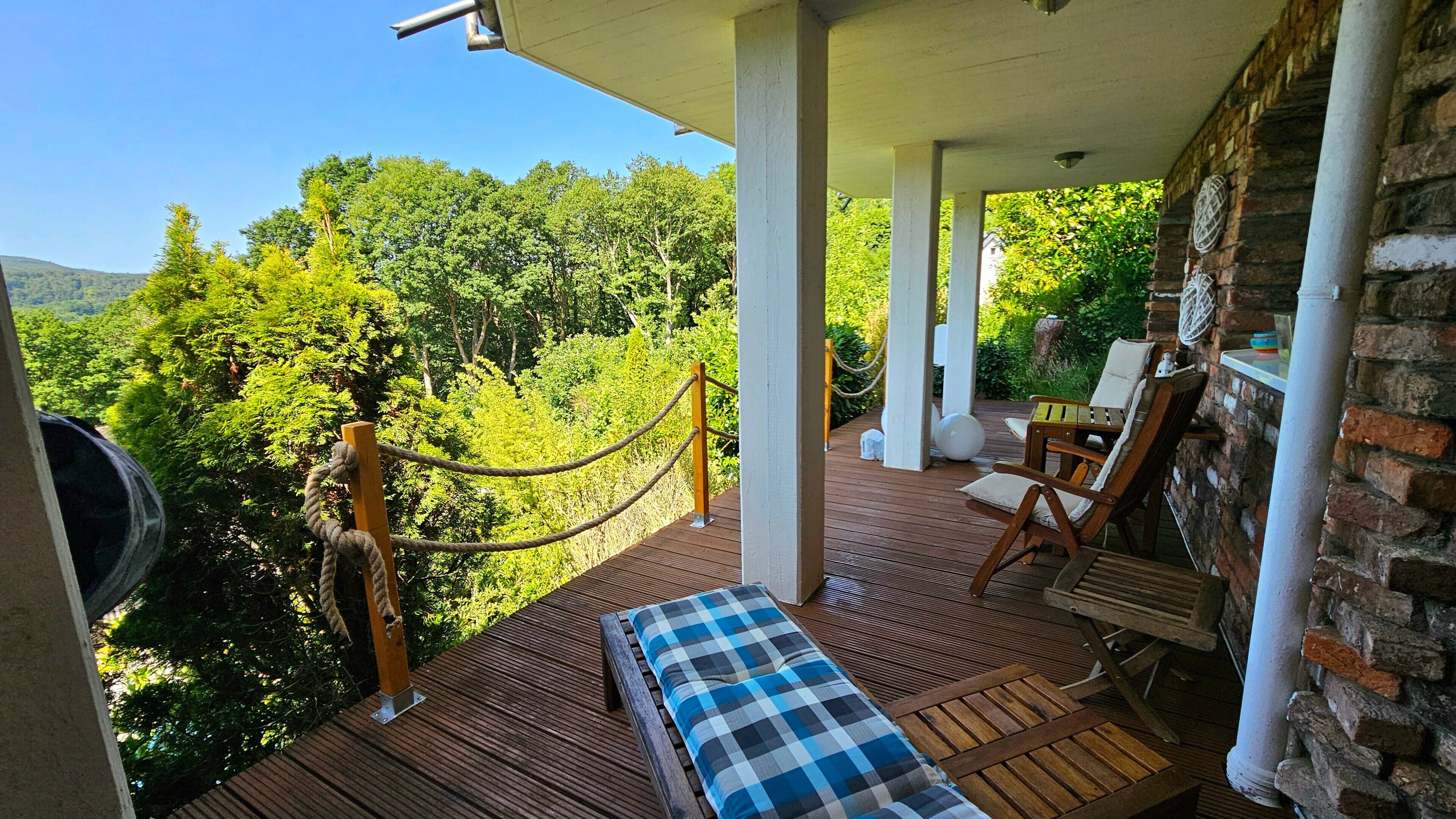 Une terrasse en bois avec un banc à carreaux, deux chaises en bois et une barrière en corde.