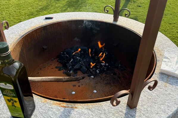 A metal outdoor grill with charcoal and flames, a bottle of olive oil, and a metal table with a handle.