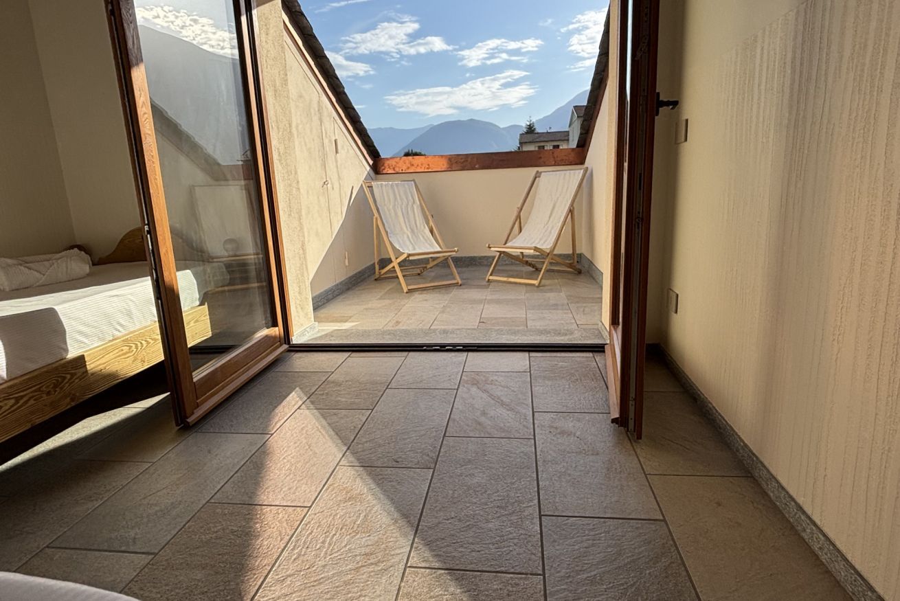 Une chambre avec un lit, une porte vitrée donnant sur un balcon équipé de deux chaises et offrant une vue sur les montagnes.