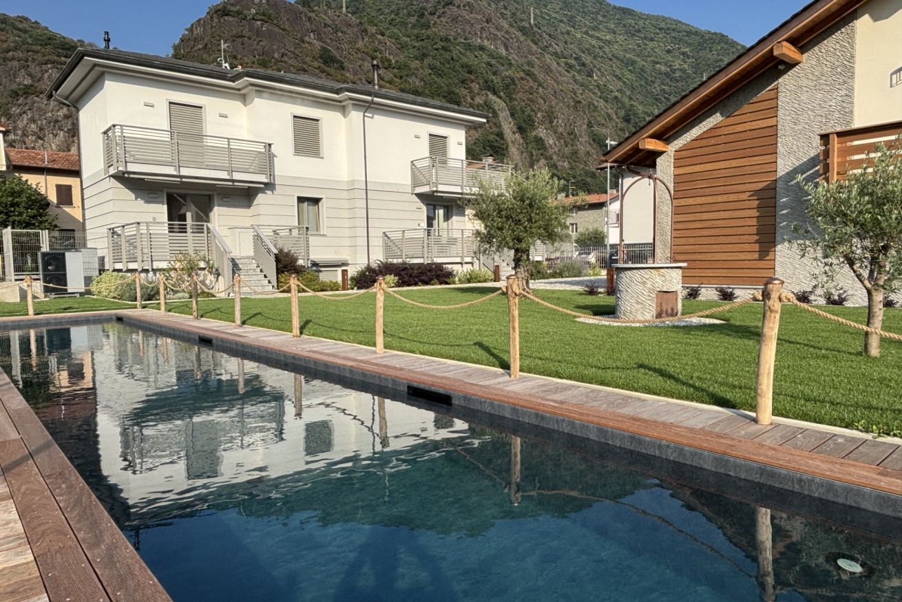 Une piscine avec une terrasse en bois, un bâtiment blanc doté de balcons et une chaîne de montagnes en arrière-plan.