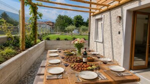 Un área de comedor al aire libre con una mesa de madera lista para una comida, rodeada por un muro de piedra y una pérgola de madera.