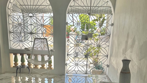 A room with two arched windows with metal grills, a white chair, a small table, and a vase on the floor.