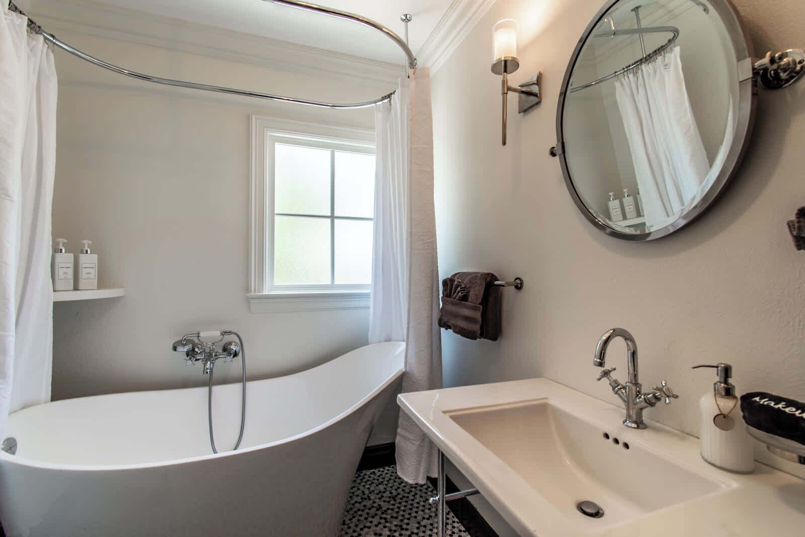 A bathroom with a freestanding bathtub, a round mirror, a sink with a handheld soap dispenser, and a window with a curtain.