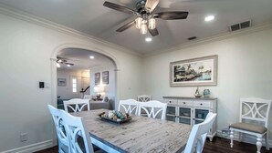 A dining room with a wooden table, white chairs, a ceiling fan, recessed lighting, a painting, and a view into another room. - Bayfront Breeze on Ocean Drive Sleeps 15 (Corpus Christi)