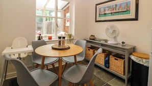 A dining area with a round table, grey chairs, a white high chair, a window with a view of a brick wall, a framed cityscape painting, a white fan, and wicker baskets on a shelf. - Robins Nest (Rhyl)