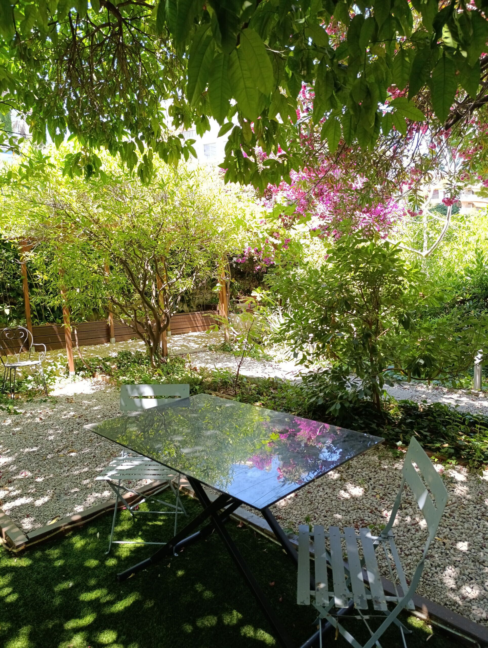 Ein Sitzbereich im Freien mit einem Glastisch und Metallstühlen, umgeben von Grünpflanzen und blühenden Blumen.