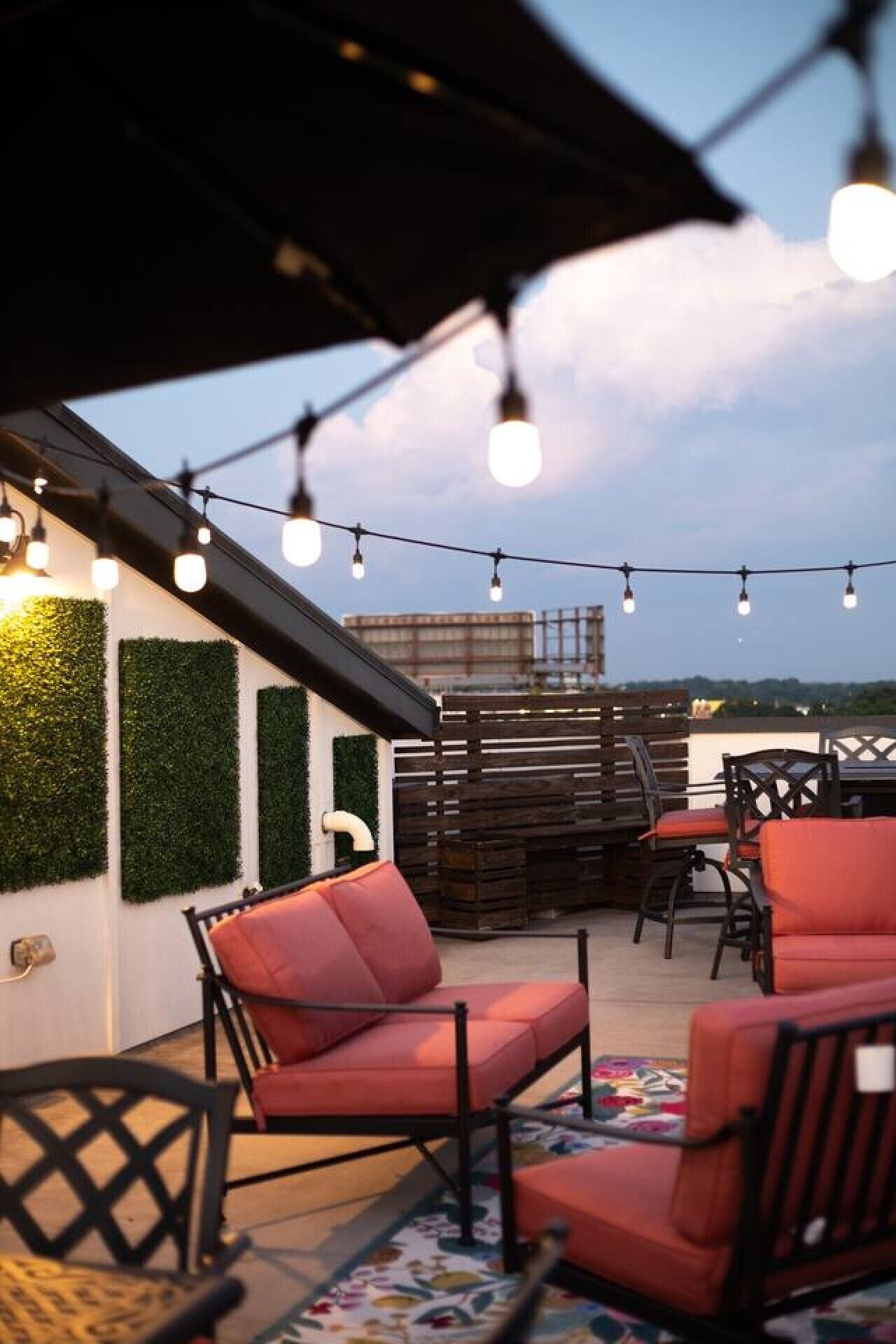 Un espacio exterior con sillas rojas y una mesa, luces colgantes y vistas del horizonte urbano.