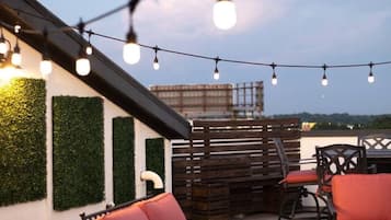 An outdoor seating area with red chairs and a table, string lights overhead, and a view of the city skyline.