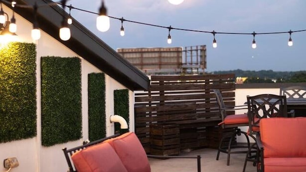 Een buitenverblijf met rode stoelen en een tafel, sfeerverlichting erboven en uitzicht op de skyline.