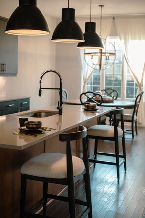 A modern kitchen with a central island, bar stools, pendant lighting, and a dining area with chairs.