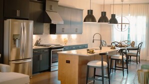 Een moderne keuken met donkere kastjes, roestvrijstalen apparatuur, hanglampen en een centraal eiland met barkrukken.
