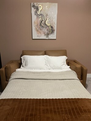 A bedroom with a bed, two pillows, a blanket, and a wall-mounted abstract painting.