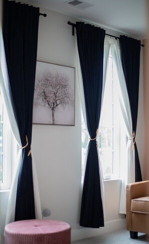 A room with two windows, each with dark curtains tied back, revealing a white wall and a framed picture of a tree. A round pink ottoman and a brown chair are visible. - Five-minute walk to downtown Atlanta with Rooftop (Atlanta)