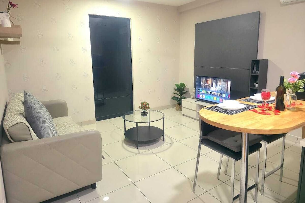 A modern living room with a sofa, a round glass coffee table, a dining table with chairs, a TV, and a small potted plant.