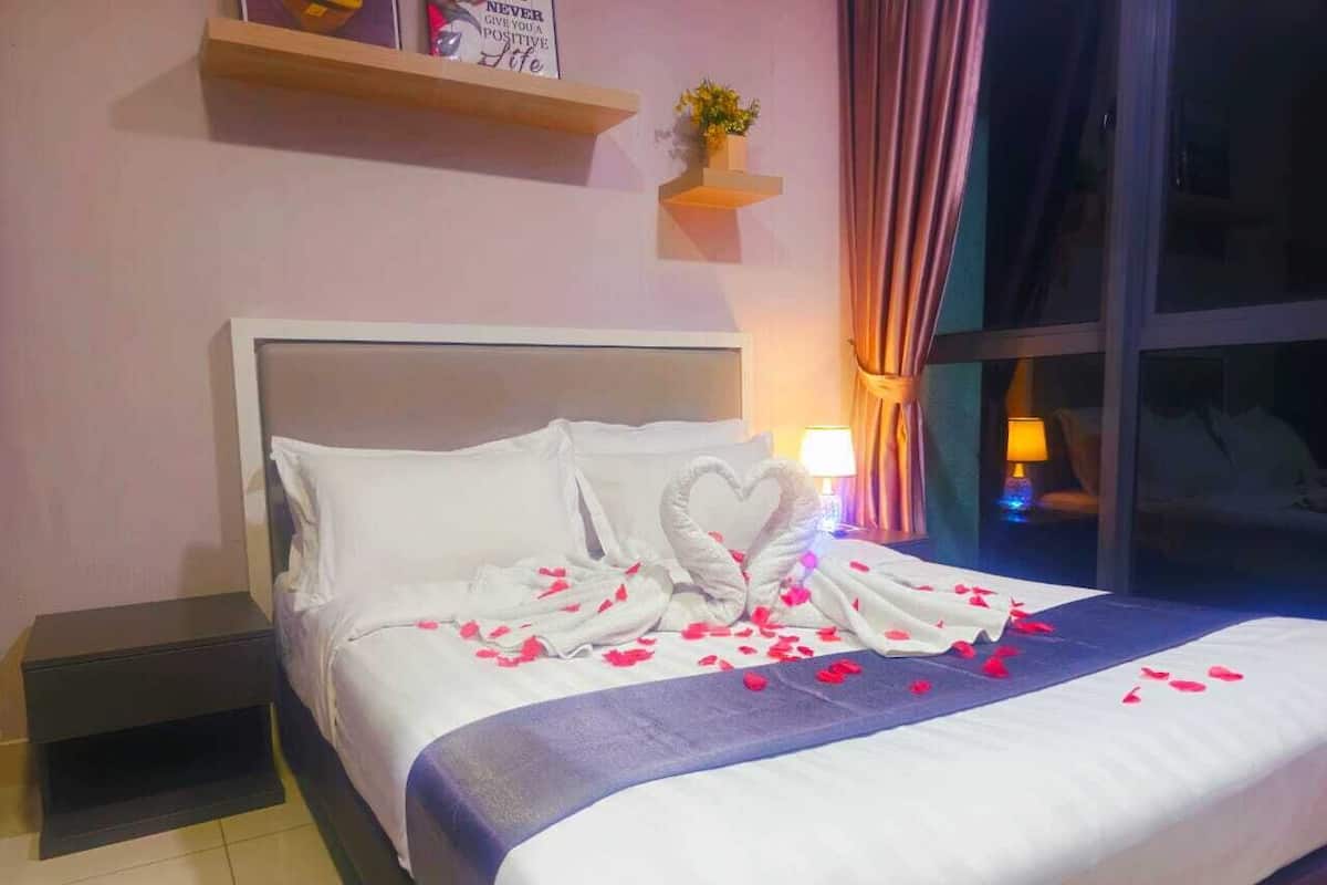 A neatly made bed with heart-shaped towels and rose petals, a bedside lamp, and a nightstand.