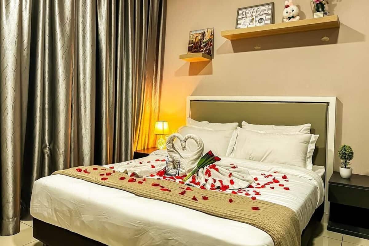 A hotel room with a bed, curtains, a nightstand, a lamp, a wall-mounted shelf with decorative items, and a small potted plant.