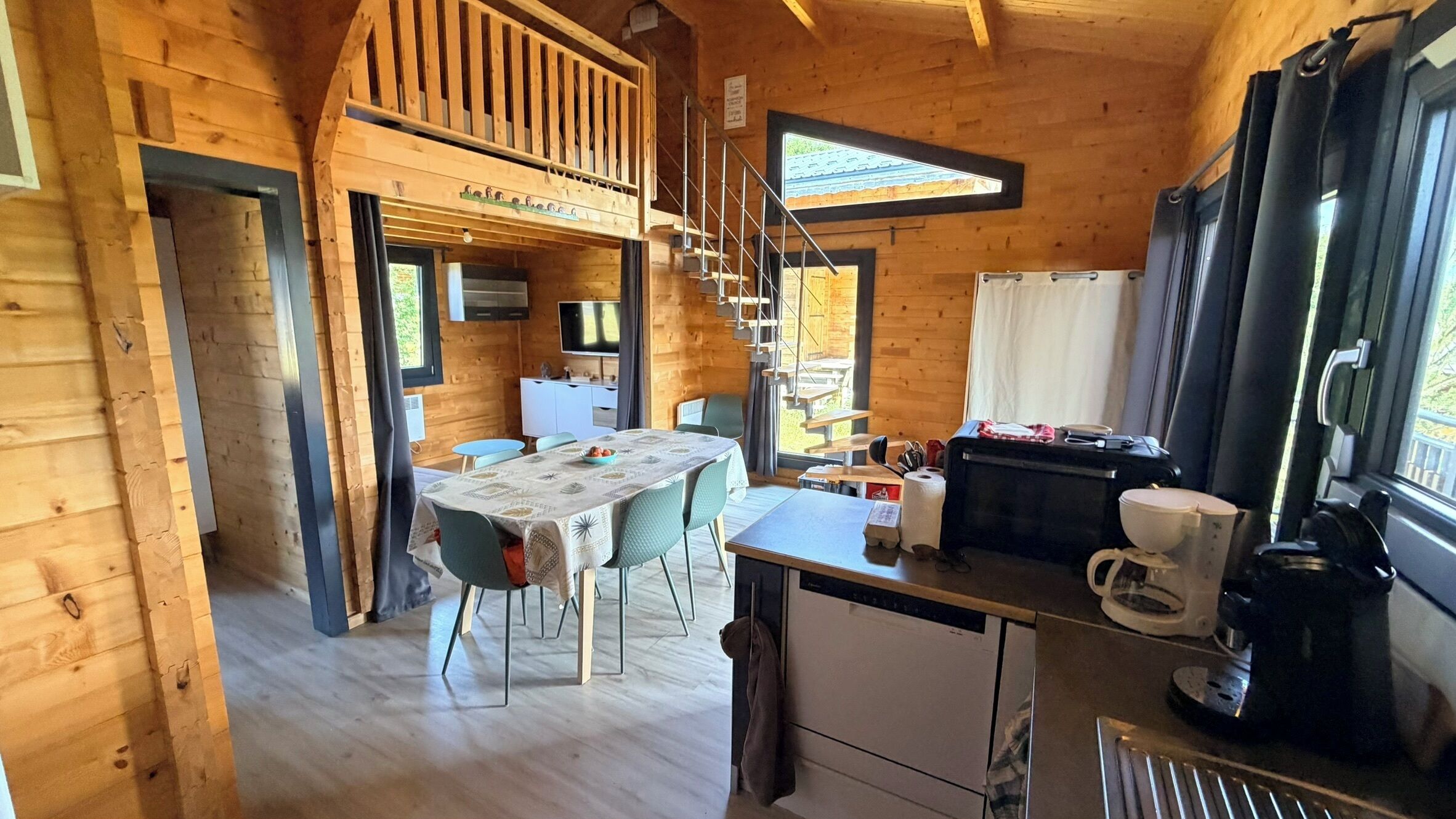 A wooden cabin interior with a dining area, kitchen, and a staircase leading to a loft.