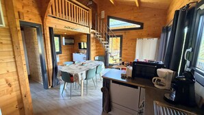 A wooden cabin interior with a dining area, kitchen, and a staircase leading to a loft. - Cozy chalet in the Haut-Jura with an indoor pool (Saint-Laurent-en-Grandvaux)