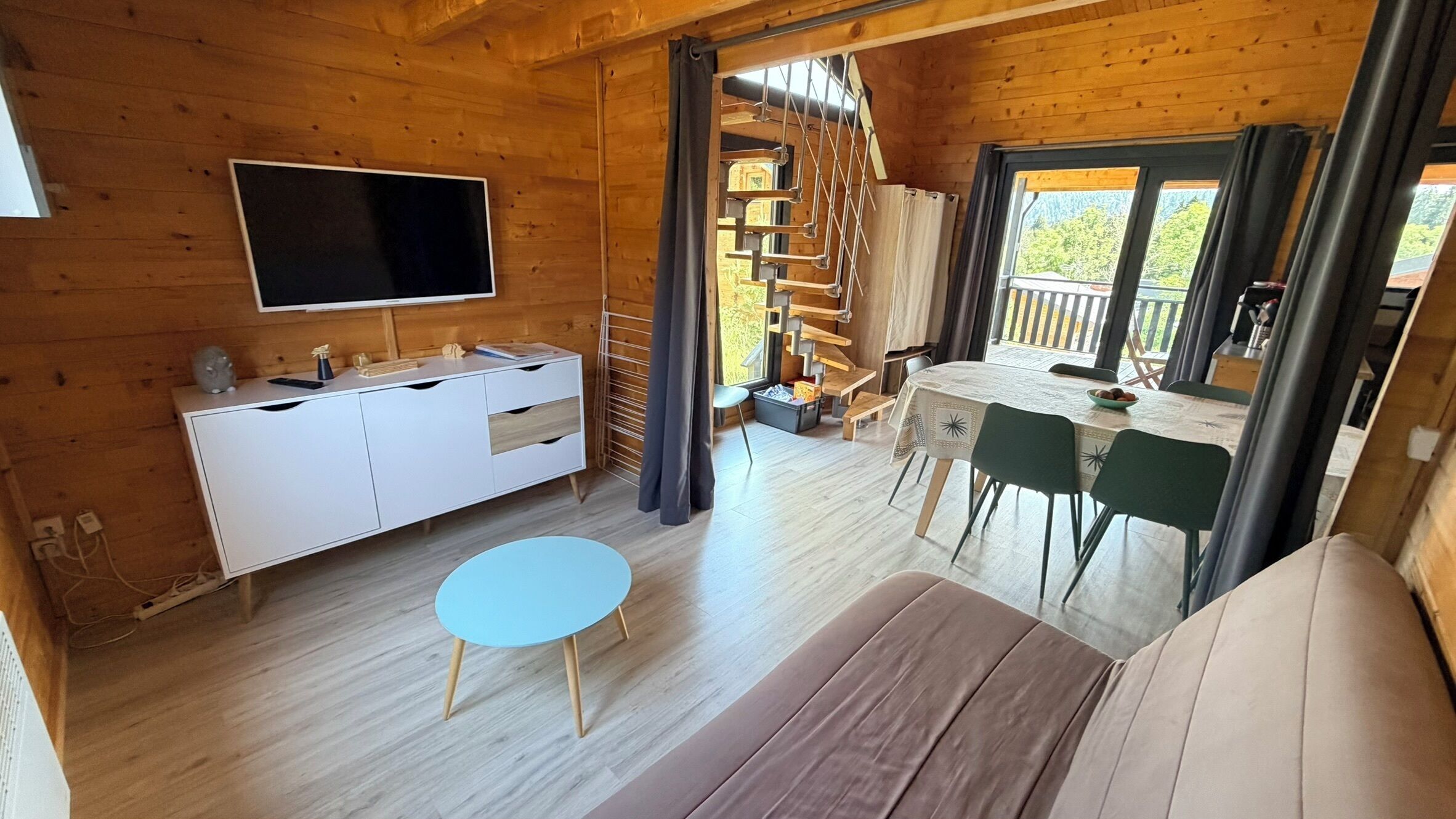 A wooden cabin interior with a TV, a dining table, chairs, a sofa, and a staircase.