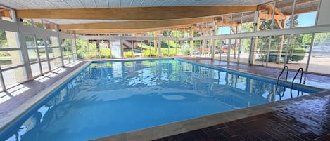 An indoor swimming pool with clear glass walls, a tiled floor, and a wooden ceiling structure.