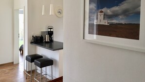 Une cuisine moderne équipée d’une machine à café, de deux tabourets de bar, d’une horloge et d’un tableau représentant un paysage, encadré.