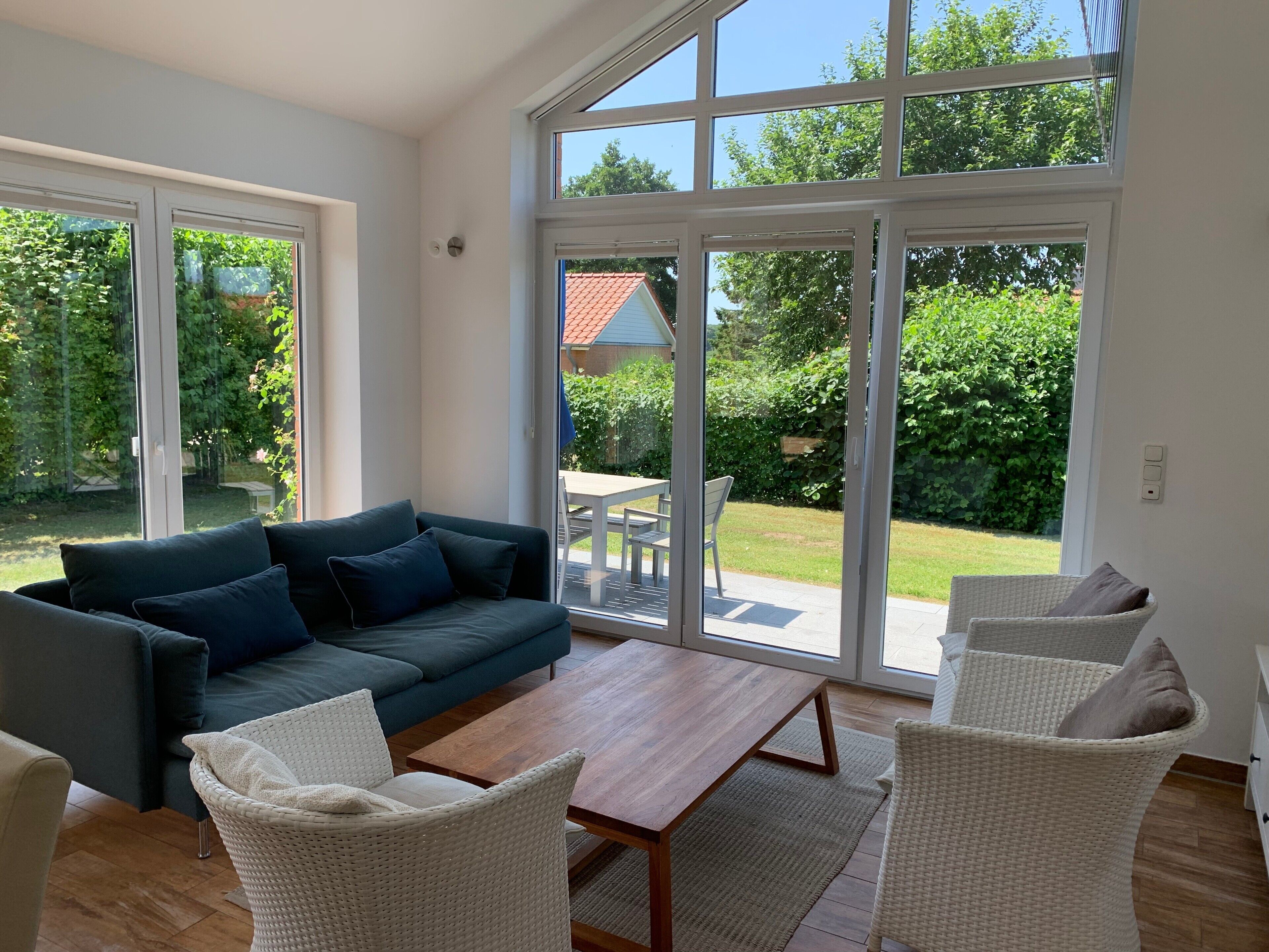 A modern living room with a sofa, coffee table, and wicker chairs, large windows, and a view of a garden.