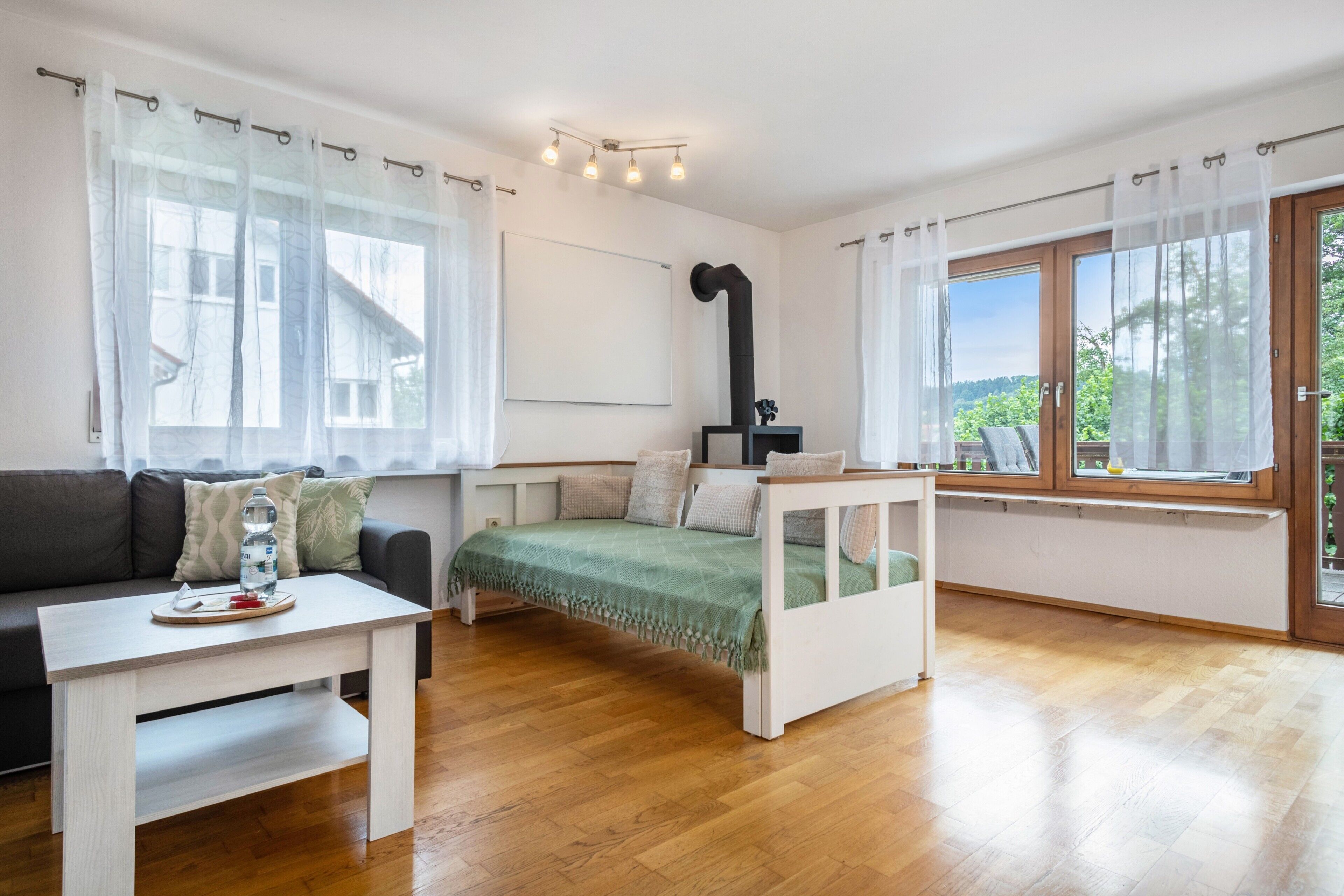 A modern, well-lit room with a bed, a sofa, a small table, and a window with a view of greenery.