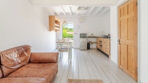 Una habitación con sofá de cuero marrón, puerta de madera, zona de cocina con refrigerador y ventana con vistas a vegetación.