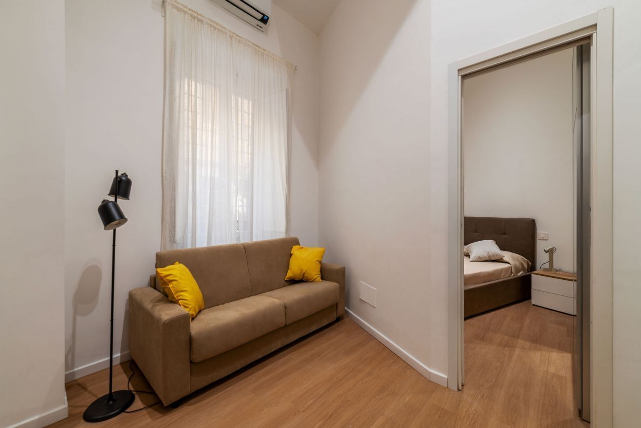 Una habitación pequeña y moderna con sofá, mesita de noche, lámpara y ventana con cortinas.