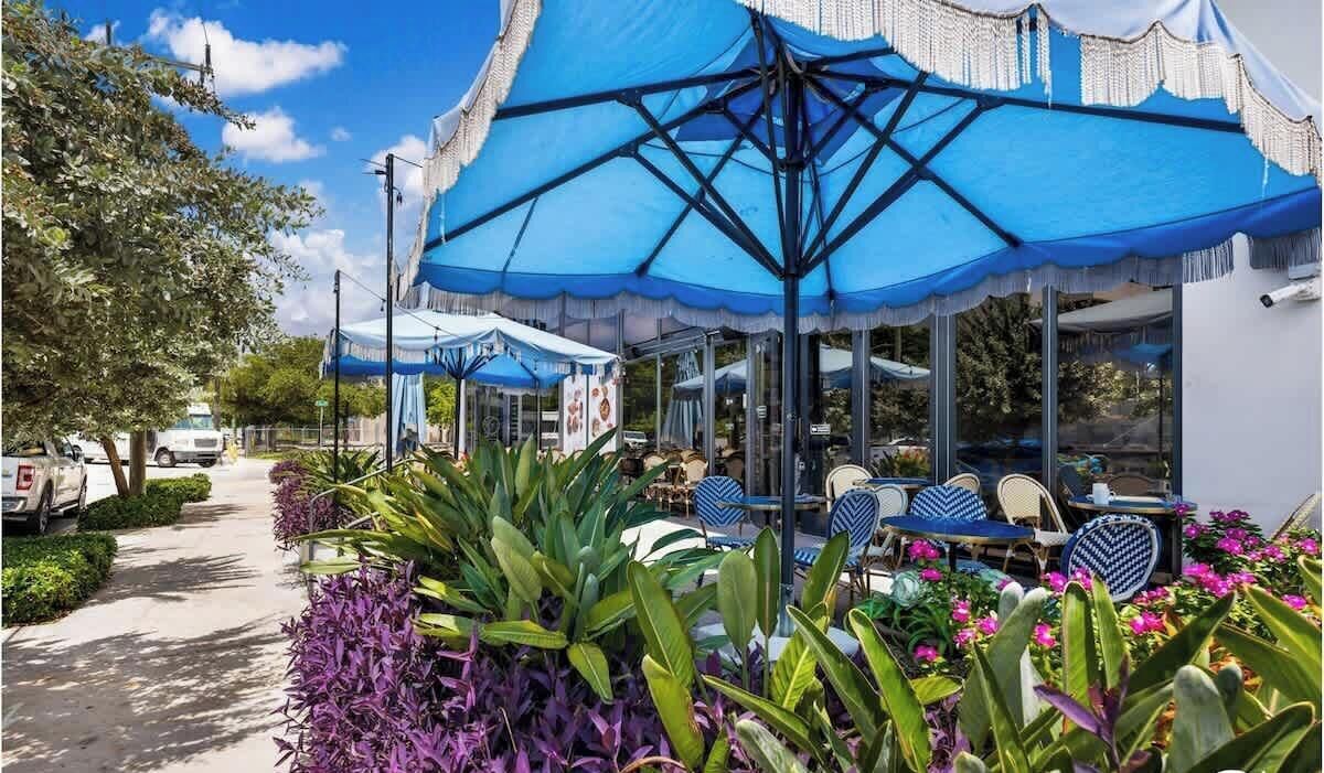 A restaurant with outdoor seating, covered by blue umbrellas, and surrounded by greenery.