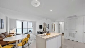 A modern kitchen with a central island, a dining area with yellow chairs, a sofa, and a large window. - High-Rise One-Bedroom Condo In Edgewater Miami (Miami)