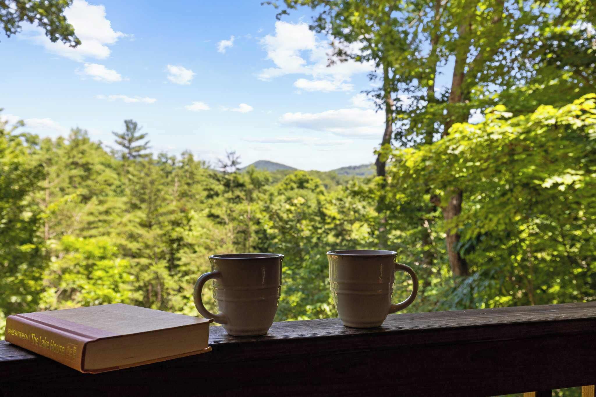 Un livre intitulé « La maison au bord du lac » et deux tasses blanches posées sur une étagère en bois, avec vue sur la forêt.