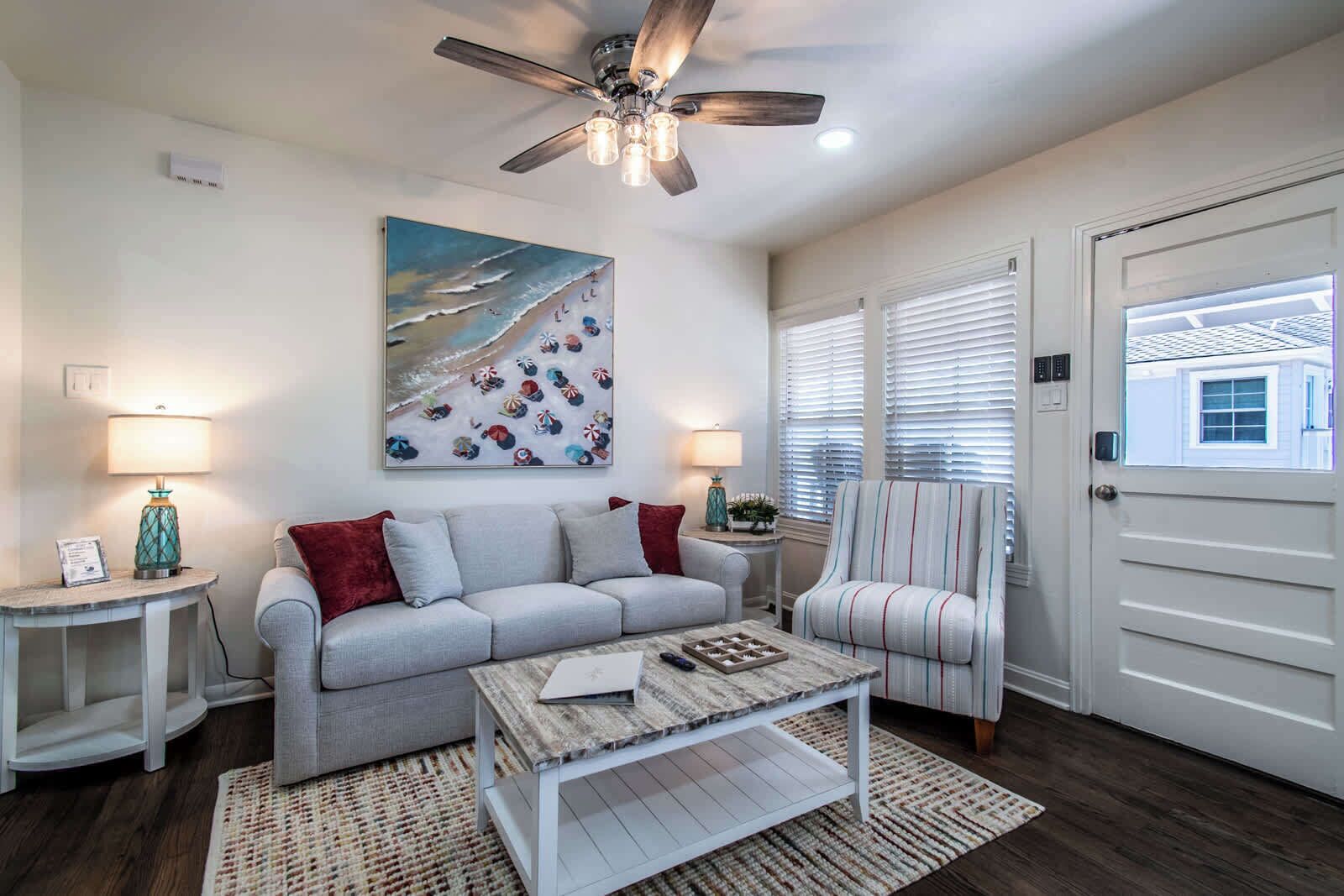 Un salon avec un canapé gris, une table basse, un ventilateur de plafond et un tableau représentant une scène de plage.