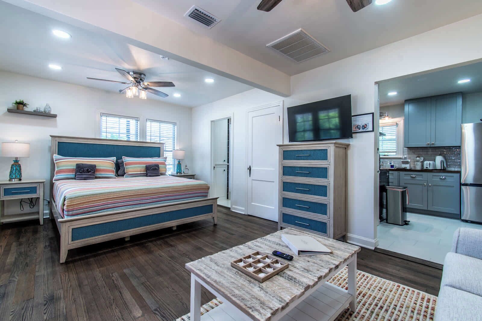 A bedroom with a bed, bedside tables, a desk, a chair, a TV, a kitchen area, and a ceiling fan.