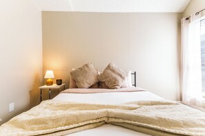 A bedroom with a bed, pillows, a bedside lamp, and a window with curtains. - Cozy 2BR Condo Near DFW Airport & Las Colinas (Irving)