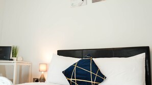 En snyggt bäddad säng med ett blått täcke, en svart sänggavel och en vit kudde med ett geometriskt mönster. Ovanför sängen hänger tre abstrakta tavlor på väggen.