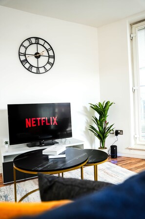 Un saló amb una televisió que mostra 'NETFLIX', un rellotge de paret, una planta en test i una taula petita.