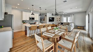 Una cocina moderna con armarios blancos, un comedor con sillas de madera y una sala de estar con sofá y televisor.