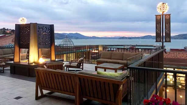 Eine Dachterrasse mit Sitzgelegenheiten im Freien, einer Glasbrüstung und Blick auf das Meer und die Berge.