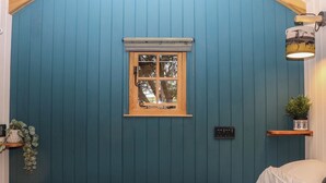 Una habitación con un muro azul con paneles, una ventana con marco de madera, una cama con colcha estampada, un estante pequeño con una planta y una lámpara fijada a la pared.