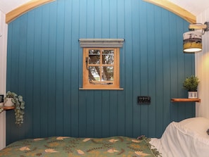 A room with a blue paneled wall, a wooden-framed window, a bed with a patterned bedspread, a small shelf with a plant, and a wall-mounted light fixture. - Goldcrest (Yelverton)