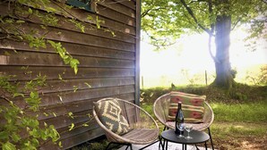 Una silla de mimbre con un cojín estampado, una pequeña mesa redonda con una botella y vasos, y vista a un área verde con un árbol y un edificio al fondo.