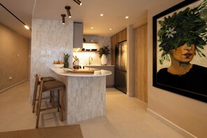 A modern kitchen with a central island, wooden stools, a refrigerator, and a framed artwork on the wall. - Two King Beds | Ocean Views | Beachfront Steps (San Juan)