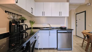 Een compacte keuken met witte kastjes, een vaatwasser, een koffieapparaat en een klein eethoekje met een stoel.