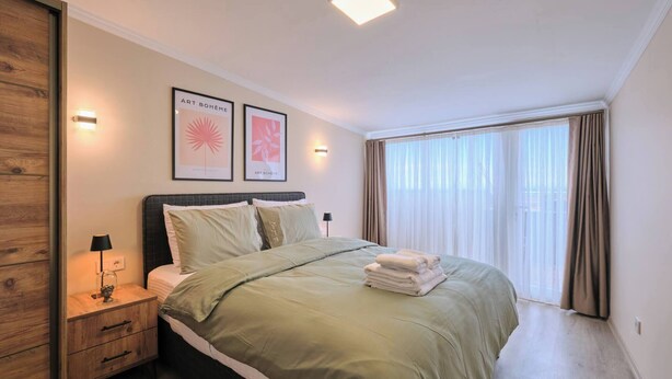 Een slaapkamer met een bed, nachtkastjes, een lamp, kunstwerken en een groot raam met gordijnen.