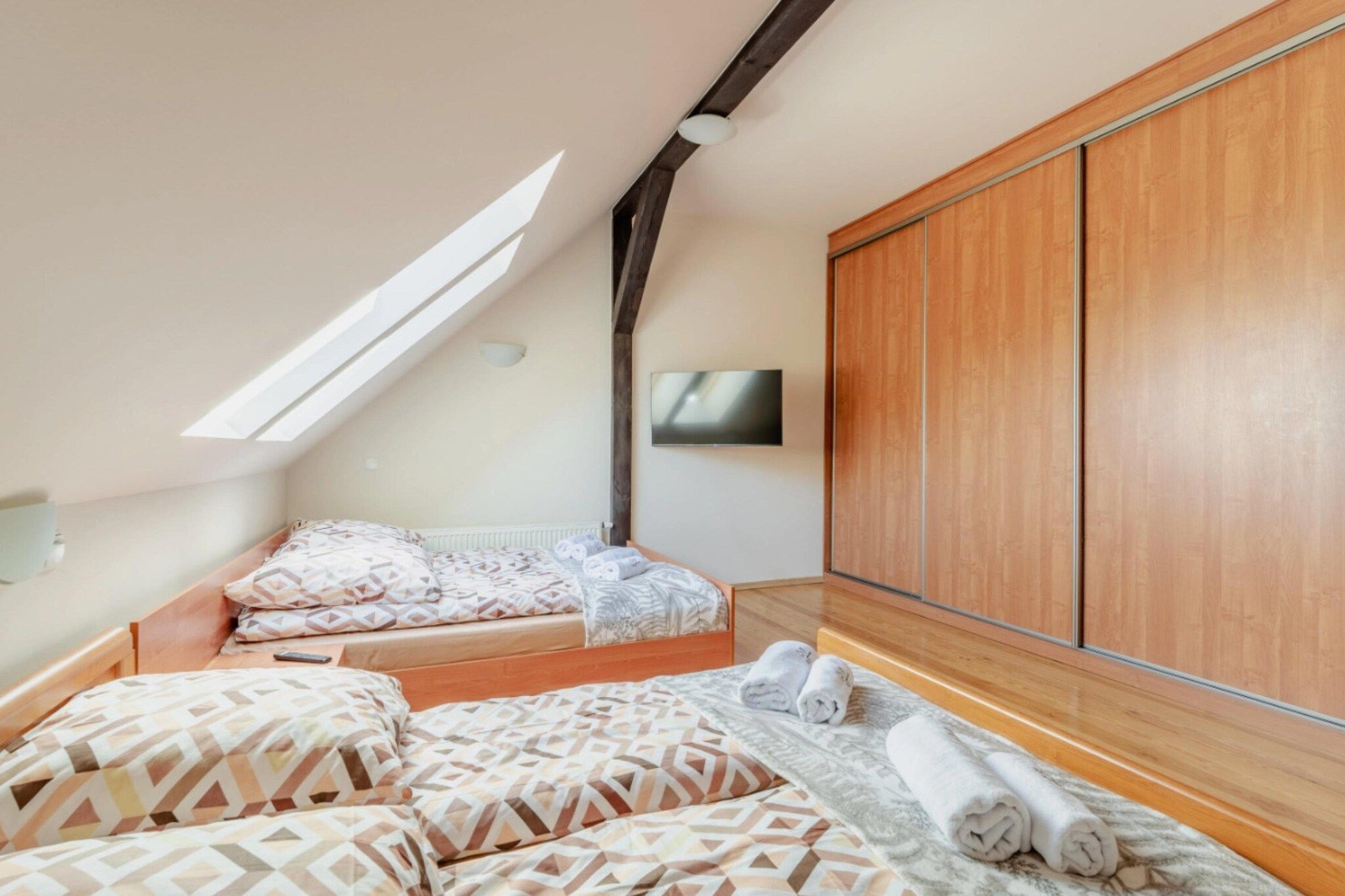A bedroom with a sloped ceiling, a bed with patterned bedding, a wooden wardrobe, and a television mounted on the wall.