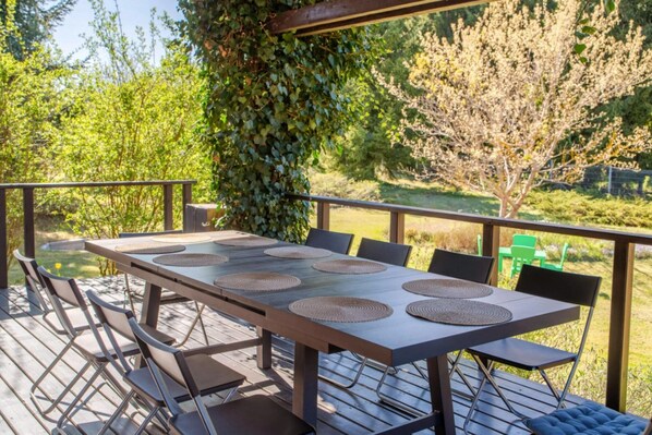 An outdoor dining area with a table and chairs, surrounded by greenery and a view of a garden.