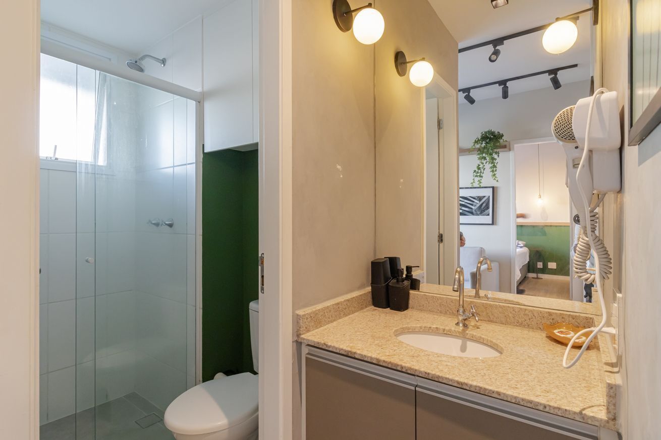 A bathroom with a glass shower enclosure, a white toilet, a beige sink with a mirror, and a hairdryer mounted on the wall.