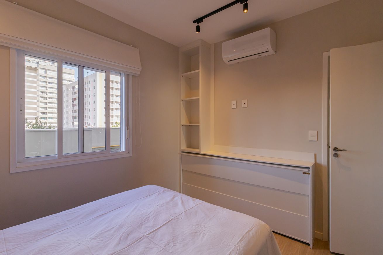 Een slaapkamer met een bed, inbouwplanken, een airconditioning en een raam met uitzicht op hoge gebouwen.